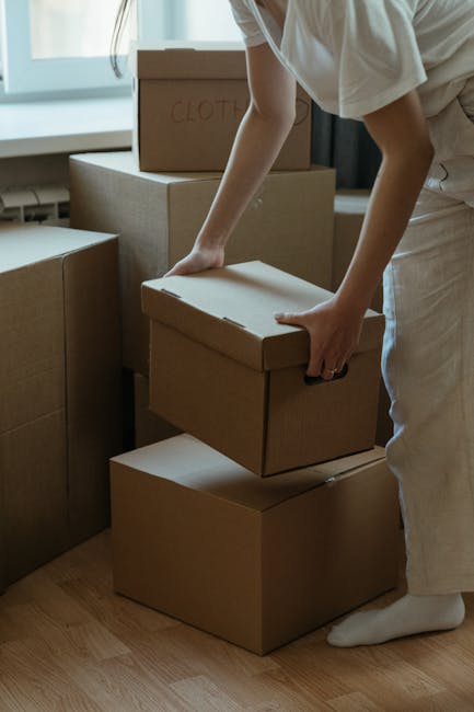 A man with dark hair and a beard, wearing a navy blue T-shirt and dark trousers, is inside a well-lit room with white walls and wooden flooring. He is holding a cardboard box, partially wrapped in cle